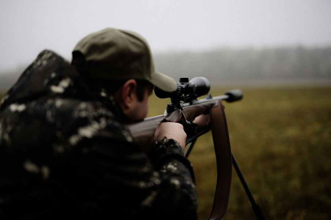 Hunter aiming a scoped bolt-action rifle from shooting sticks in an open field